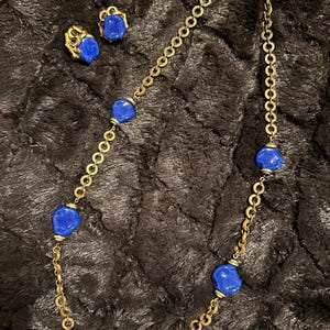 May include: A gold-toned chain necklace featuring five irregular, smooth, deep blue glass beads spaced along the links, accompanied by a pair of matching clip-on earrings, each displaying a single blue glass bead and small gold-toned accents.