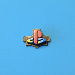 May include: A bronze-coloured PlayStation logo pin. The logo features a red 'P' shape above a yellow, blue, and black design. The pin is set against a solid blue background.