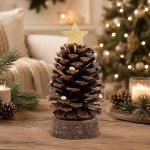 Pode incluir: Uma pinha decorativa em forma de árvore de Natal, com uma estrela de madeira dourada no topo e pequenas contas douradas entre as escamas. A pinha está montada sobre uma base circular de madeira natural com casca visível nas bordas.