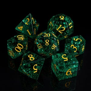 May include: A set of seven polyhedral dice in shades of green with gold numbering. The dice are made of a translucent material with a textured, crackled appearance. The dice are used for tabletop role-playing games.