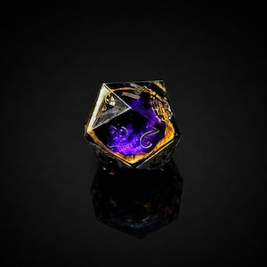 K&ouml;nnte beinhalten: Ein D20-W&uuml;rfel aus klarem Harz mit einem violett-goldenen Inneren. Der W&uuml;rfel hat goldene Zahlen und ist vor einem schwarzen Hintergrund platziert. Die Reflexion des W&uuml;rfels ist auf der Oberfl&auml;che sichtbar. Ein beliebtes Objekt f&uuml;r Tabletop-Spiele.