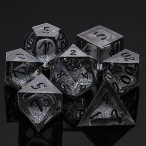 May include: A set of seven clear, resin dice with black numbering and embedded silver glitter. The dice are various geometric shapes, including a d4, d6, d8, d10, d12, d20, and a percentile die. The dice are for tabletop gaming.