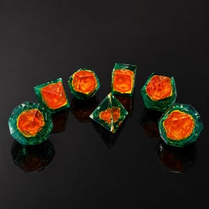 Puede incluir: Un juego de siete dados poliédricos con una capa exterior de color verde esmeralda translúcido y un núcleo naranja vibrante y texturizado. Cada dado muestra números de color dorado grabados en la superficie con formas geométricas.