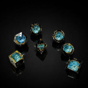 May include: A collection of seven polyhedral dice with a gemstone aesthetic. Each die features a translucent body with a vibrant blue interior, accented by gold-colored trim. The dice are arranged on a reflective black surface.