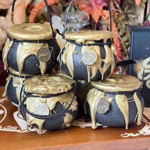 May include: Four black cauldron-shaped containers with gold-colored lids and dripping gold paint accents. Each container features a black handle and a small circular metal charm hanging from a black cord, displaying the zodiac signs Aries, Cancer, Sagittarius, and Taurus.