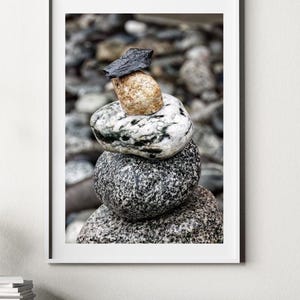 May include: A framed vertical wall art print featuring a stack of five smooth, rounded stones balanced on top of each other. The stones vary in color and texture, ranging from dark grey granite and speckled white to a smooth tan and a sharp black top stone.