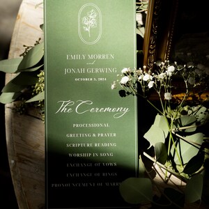 Op de afbeelding: Een rechthoekige donkergroene trouwceremonie programmakaart met een wit lijntekening bloemenembleem, de namen Emily Morren en Jonah Gerwing, de datum 3 oktober 2024 en een lijst met ceremonie-evenementen gedrukt in witte serif- en scriptlettertypes.