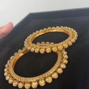 May include: Two gold bangles with a textured design. The bangles feature small, round, beaded embellishments around the outer edge. The jewellery is displayed on a black surface.