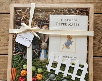 May include: A wooden box gift set featuring a copy of "The Tale of Peter Rabbit" book, a wooden Peter Rabbit figurine, and miniature vegetables. The set includes a white picket fence and is tied with a cream ribbon.