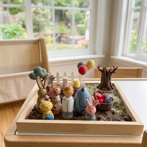 May include: A wooden tray filled with miniature figurines of cartoon characters, including a bear, tiger, rabbit, and pig. The tray also contains a white picket fence, a tree, and a basket of fruit. The scene is set on a wooden table.