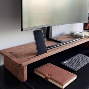Peut inclure: Un support d'écran en bois avec une finition brun foncé et une fente intégrée pour smartphone. Le support présente un design en contreplaqué stratifié sur les bords et soutient un écran d'ordinateur. Un carnet brun et un clavier noir sont posés.