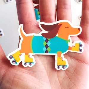 May include: A hand holding a sticker of an orange dachshund wearing a blue and green sweater and roller skates. The dachshund is on a white background with other dachshund stickers.