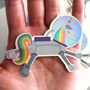May include: A silver robot unicorn sticker with a rainbow mane and tail. The unicorn is wearing a space helmet with a blue and white galaxy design. The sticker is on a white background.