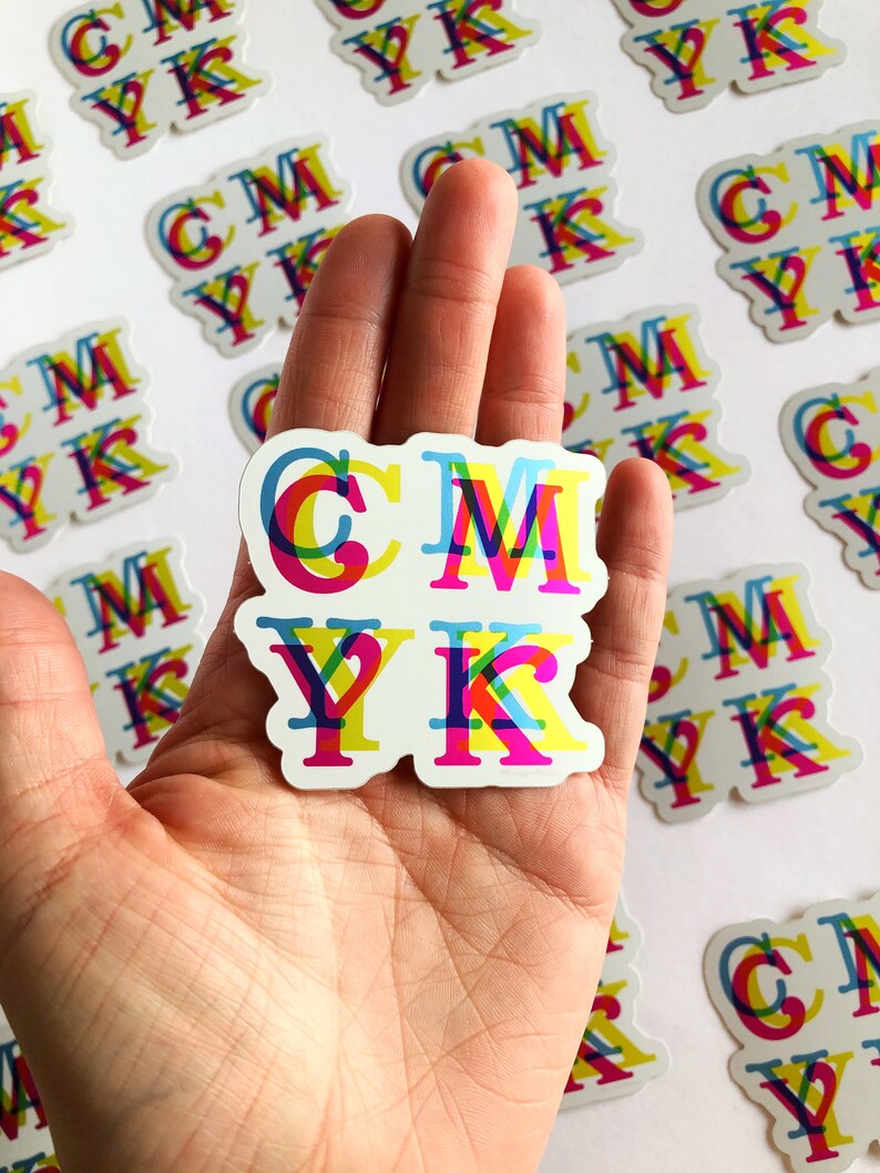 May include: A hand holding a sticker with the letters CMYK in a colorful, layered design. The letters are outlined in black and each letter is made up of overlapping colors, creating a layered effect. The sticker is on a white background.
