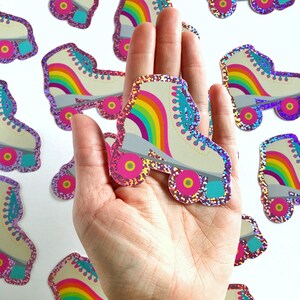 May include: A hand holding a rainbow roller skate sticker with glitter. The sticker is white with a rainbow design and turquoise wheels.