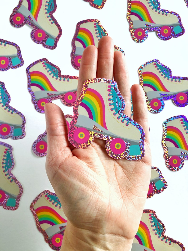 May include: A hand holding a rainbow roller skate sticker with glitter. The sticker is white with a rainbow design and turquoise wheels.