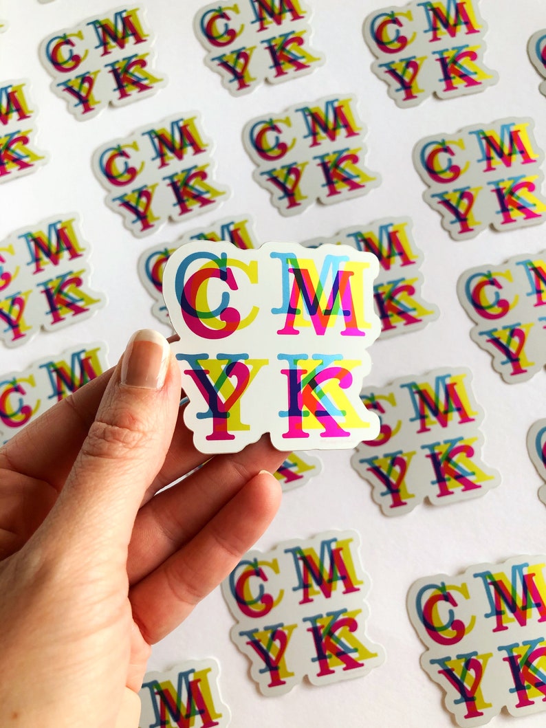 May include: A close-up of a hand holding a sticker with the letters CMYK printed in overlapping colors. The letters are printed in a rainbow pattern of cyan, magenta, yellow, and black.
