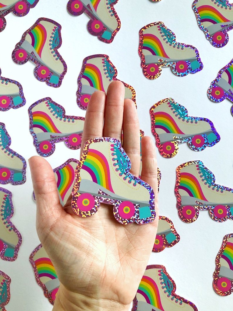 May include: A hand holding a white roller skate with rainbow stripes and pink glitter. The skate has turquoise wheels and pink accents.