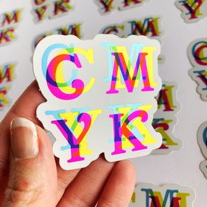 May include: A close-up of a sticker with the letters CMYK printed in a rainbow color scheme. The letters are layered on top of each other, creating a 3D effect.