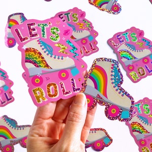 May include: A set of pink and white roller skate stickers with rainbow accents. The stickers feature the text "Let's Roll" and glitter accents.
