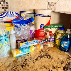 May include: A collection of household cleaning and dining supplies, including paper towels, plastic cups, plates, cutlery, dish soap, and sponges. The items are arranged on a countertop with a patterned backsplash.