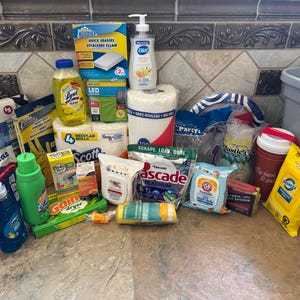 May include: A selection of household cleaning and personal care products, such as paper towels, dish soap, and cleaning wipes. The items are arranged on a tiled surface, with a gray storage bin in the background. Various brands and product types are visible.