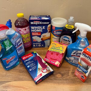 May include: A collection of household cleaning supplies including Windex, Lysol, Pine-Sol, Mr. Clean Magic Eraser, Clorox wipes, Dawn dish soap, a Scrub Mommy sponge, Playtex Living gloves, and Hefty trash bags arranged on a wooden surface.
