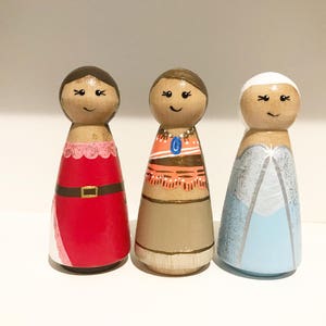 K&ouml;nnte beinhalten: Drei handbemalte Holzpuppen. Eine Puppe tr&auml;gt ein rotes Kleid mit goldenem G&uuml;rtel, eine andere ein beiges Kleid mit orangefarbenem Oberteil und die dritte ein blaues Kleid mit silbernem Design. Jede Puppe hat ein l&auml;chelndes Gesicht.