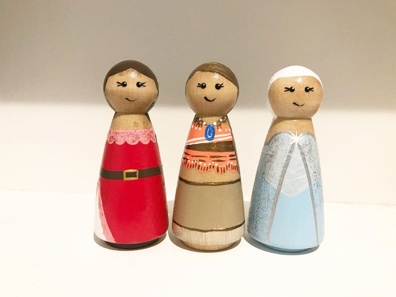 K&ouml;nnte beinhalten: Drei handbemalte Holzpuppen. Eine Puppe tr&auml;gt ein rotes Kleid mit goldenem G&uuml;rtel, eine andere ein beiges Kleid mit orangefarbenem Oberteil und die dritte ein blaues Kleid mit silbernem Design. Jede Puppe hat ein l&auml;chelndes Gesicht.