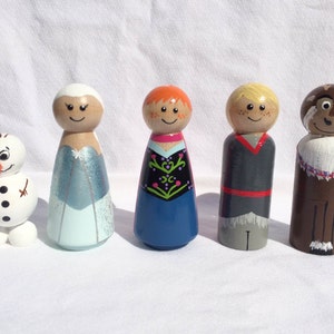 May include: A collection of five wooden peg dolls featuring characters from a popular animated film. The dolls include a snowman, two female characters in blue and red dresses, a male character in a gray outfit, and a brown reindeer.