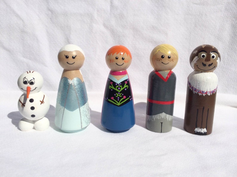 May include: A collection of five wooden peg dolls featuring characters from a popular animated film. The dolls include a snowman, two female characters in blue and red dresses, a male character in a gray outfit, and a brown reindeer.