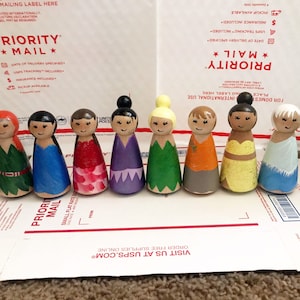 May include: A collection of eight hand-painted wooden peg dolls, each with a unique hairstyle and dress. The dolls feature various colors, including red, blue, pink, purple, green, orange, and yellow. They are arranged on a cardboard surface.