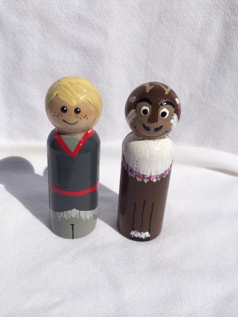May include: Two hand-painted wooden peg dolls. One doll has blonde hair, a gray and red outfit, and freckles. The other doll has brown hair, a white and purple collar, and a brown outfit. Both dolls have smiling faces.