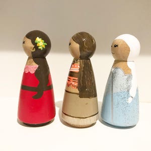 K&ouml;nnte beinhalten: Drei handbemalte Holzpuppen. Die Puppen sind bemalt, um Figuren darzustellen, mit Details wie Kleidung und Haaren. Die erste Puppe tr&auml;gt ein rotes Kleid, die zweite ein beiges Outfit und die dritte ein blaues Kleid.