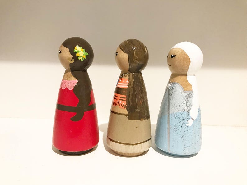 K&ouml;nnte beinhalten: Drei handbemalte Holzpuppen. Die Puppen sind bemalt, um Figuren darzustellen, mit Details wie Kleidung und Haaren. Die erste Puppe tr&auml;gt ein rotes Kleid, die zweite ein beiges Outfit und die dritte ein blaues Kleid.