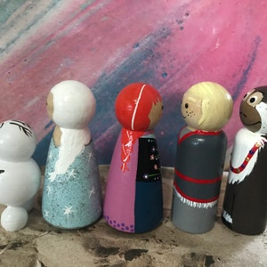 May include: A collection of five hand-painted wooden peg dolls. The dolls feature various characters with detailed facial features and clothing. The color palette includes white, blue, red, brown, and gray. The dolls are arranged in a row, showcasing their unique designs.