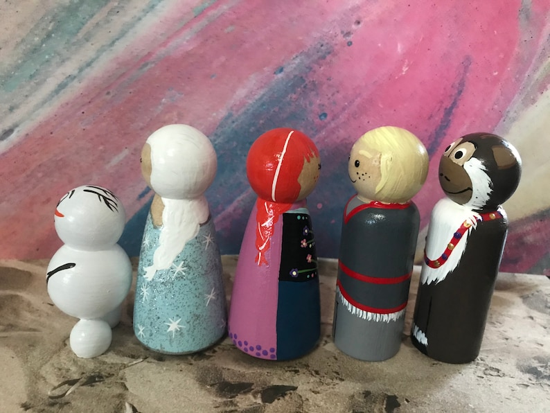 May include: A collection of five hand-painted wooden peg dolls. The dolls feature various characters with detailed facial features and clothing. The color palette includes white, blue, red, brown, and gray. The dolls are arranged in a row, showcasing their unique designs.