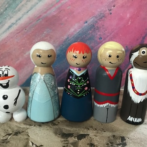 May include: A collection of five hand-painted wooden peg dolls. The dolls include a snowman, a princess in a light blue dress, a girl with red hair, a boy with blonde hair, and a monkey. Each doll has a unique design and facial expression.