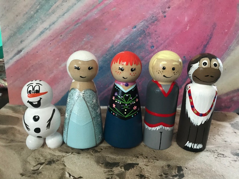 May include: A collection of five hand-painted wooden peg dolls. The dolls include a snowman, a princess in a light blue dress, a girl with red hair, a boy with blonde hair, and a monkey. Each doll has a unique design and facial expression.