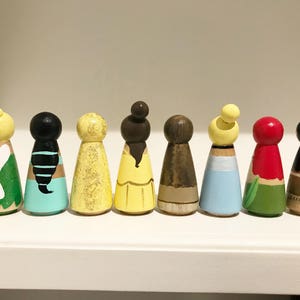 K&ouml;nnte beinhalten: Eine Sammlung von acht bemalten Holzpuppen, jede mit einzigartigen Farben und Designs. Die Puppen weisen verschiedene Frisuren und Outfits auf, darunter Gelb, Schwarz und Rot. Sie sind auf einem wei&szlig;en Regal angeordnet und pr&auml;sentieren eine handwerkliche &Auml;sthetik.