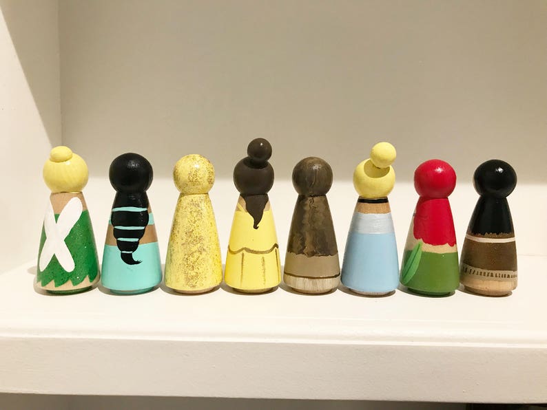 K&ouml;nnte beinhalten: Eine Sammlung von acht bemalten Holzpuppen, jede mit einzigartigen Farben und Designs. Die Puppen weisen verschiedene Frisuren und Outfits auf, darunter Gelb, Schwarz und Rot. Sie sind auf einem wei&szlig;en Regal angeordnet und pr&auml;sentieren eine handwerkliche &Auml;sthetik.