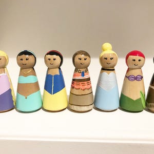 K&ouml;nnte beinhalten: Sieben handbemalte Holzpuppen, die jeweils eine andere Prinzessin darstellen. Die Puppen haben bemalte Gesichter und sind in farbenfrohen Outfits gekleidet, darunter Kleider. Die Puppen sind in einer Reihe auf einem wei&szlig;en Regal angeordnet.