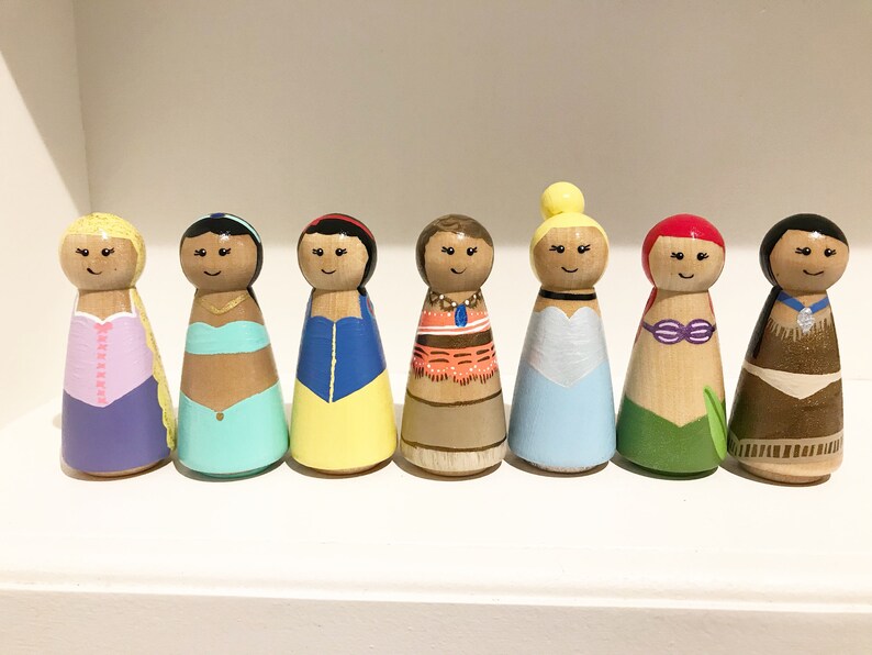 K&ouml;nnte beinhalten: Sieben handbemalte Holzpuppen, die jeweils eine andere Prinzessin darstellen. Die Puppen haben bemalte Gesichter und sind in farbenfrohen Outfits gekleidet, darunter Kleider. Die Puppen sind in einer Reihe auf einem wei&szlig;en Regal angeordnet.