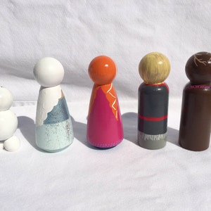 May include: A collection of five hand-painted wooden peg dolls. The dolls feature various designs, including a snowman, a figure with a blue and white dress, and characters with orange, blonde, and brown hair. The dolls are painted with bright colors and are standing on a white surface.