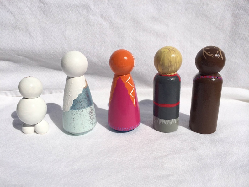 May include: A collection of five hand-painted wooden peg dolls. The dolls feature various designs, including a snowman, a figure with a blue and white dress, and characters with orange, blonde, and brown hair. The dolls are painted with bright colors and are standing on a white surface.