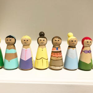 K&ouml;nnte beinhalten: Acht Holzpuppen, die jeweils eine Prinzessin darstellen. Die Puppen haben detaillierte Kleidung und Frisuren mit einem l&auml;chelnden Gesicht. Die Puppen werden auf einem wei&szlig;en Regal ausgestellt.