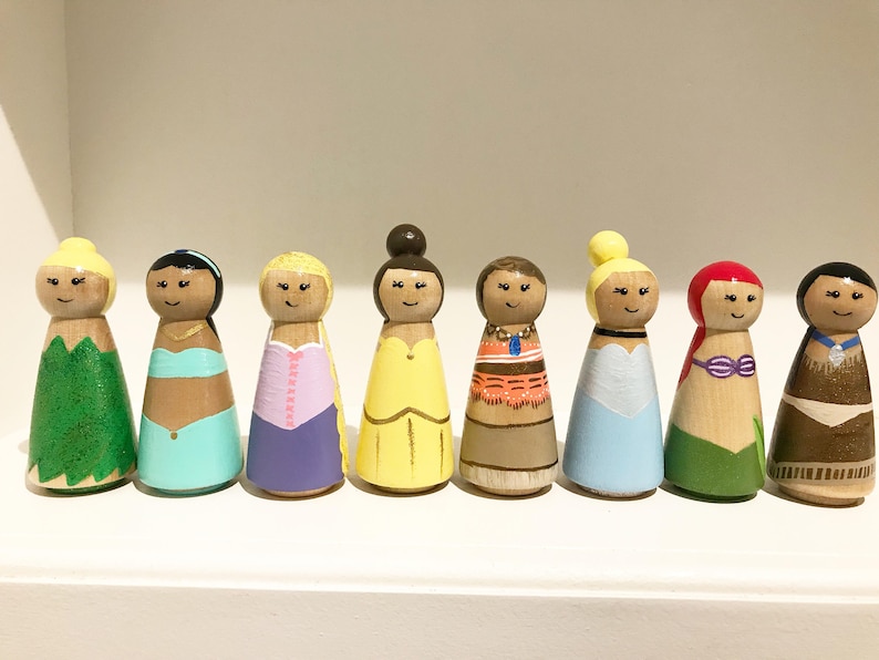 K&ouml;nnte beinhalten: Acht Holzpuppen, die jeweils eine Prinzessin darstellen. Die Puppen haben detaillierte Kleidung und Frisuren mit einem l&auml;chelnden Gesicht. Die Puppen werden auf einem wei&szlig;en Regal ausgestellt.