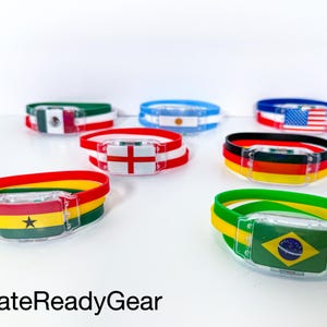 May include: A collection of silicone wristbands featuring various national flags, including Mexico, Argentina, the United States, England, Germany, Ghana, and Brazil. Each band has a clear plastic rectangular clasp. The text GateReadyGear is visible.