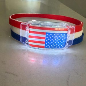 May include: A flexible silicone wristband featuring three stacked layers in red, white, and blue. A clear plastic rectangular housing is attached to the front, displaying a printed American flag graphic with red stripes and a blue field with white stars.