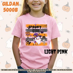 Puede incluir: Una camiseta de manga corta de color rosa claro con un estampado gráfico que incluye el texto "SPOOKY GIRL" y "HALLOWEEN PARTY". El diseño muestra un fantasma con un sombrero de bruja morado, un gato negro, un esqueleto, una telaraña y un caldero.